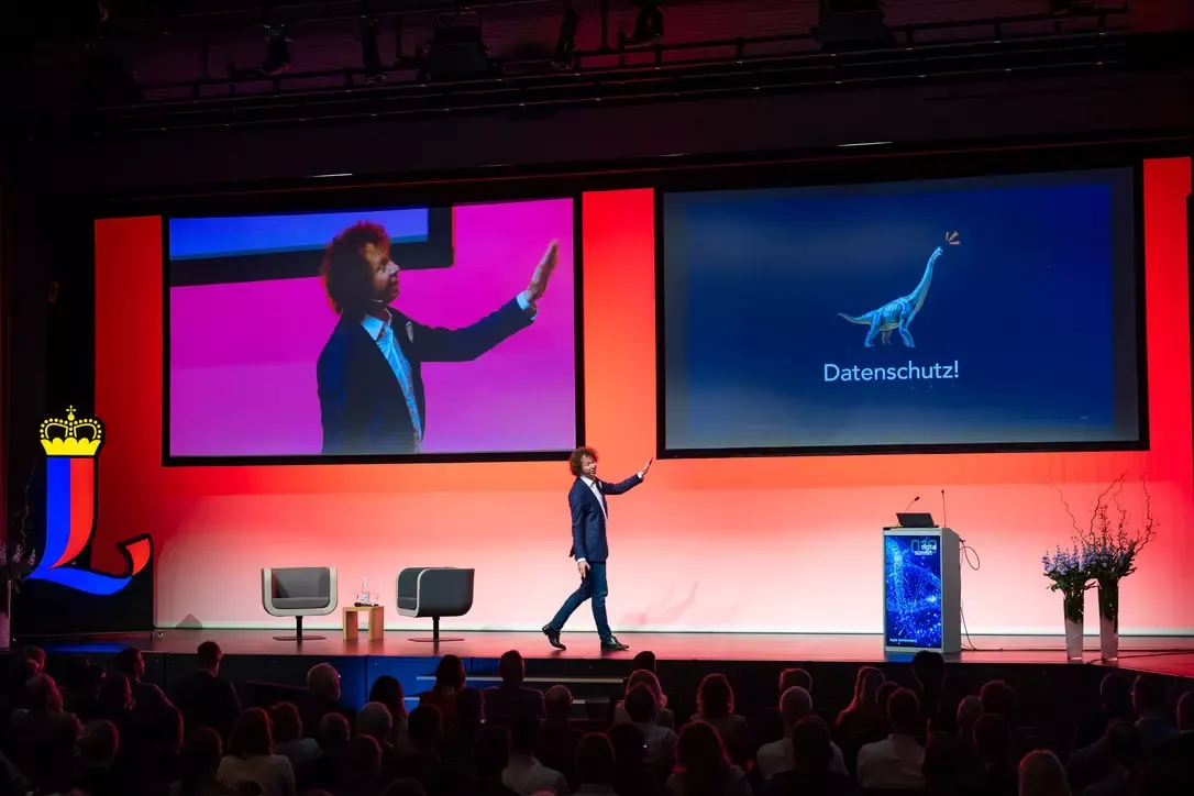 Speaker in a suit gesturing towards a presentation screen displaying a dinosaur illustration and the word 'Datenschutz!' with an audience watching.