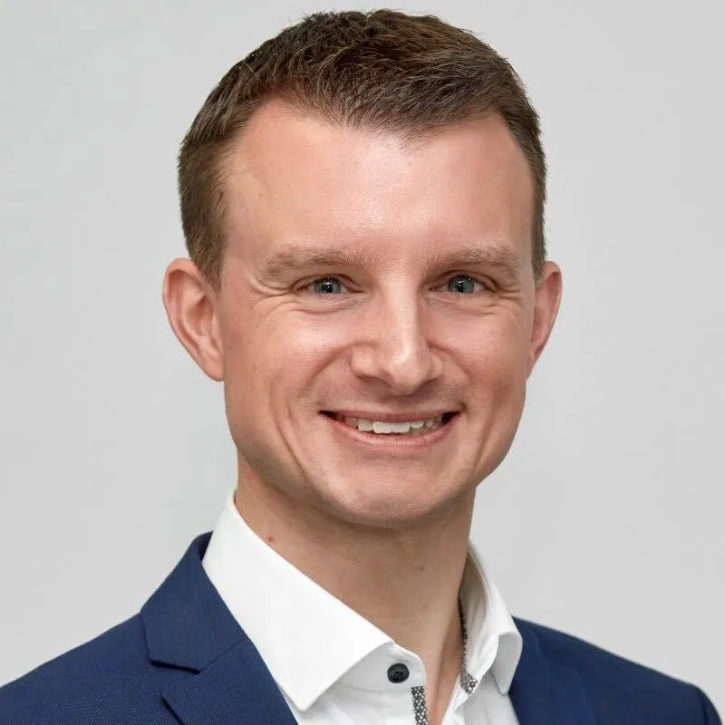 Smiling man with short brown hair wearing a blue blazer and white shirt against light gray background.