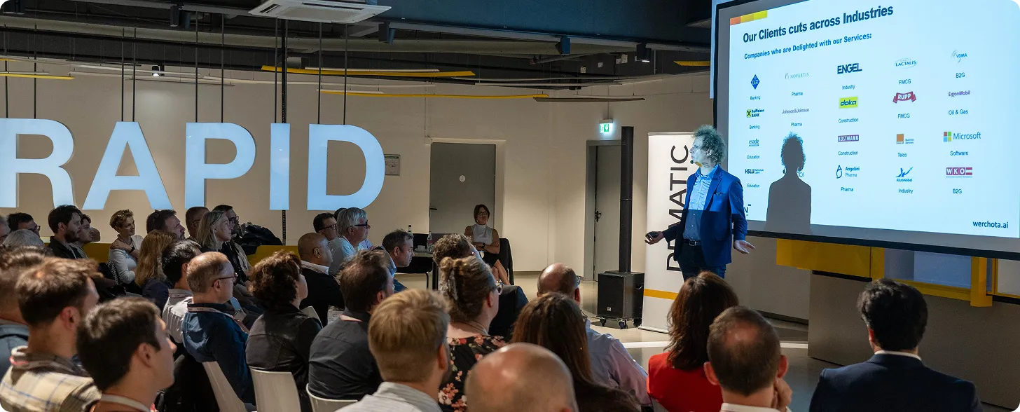 Presenter in blue suit giving a presentation to an attentive audience in a conference room with a large screen displaying client logos.