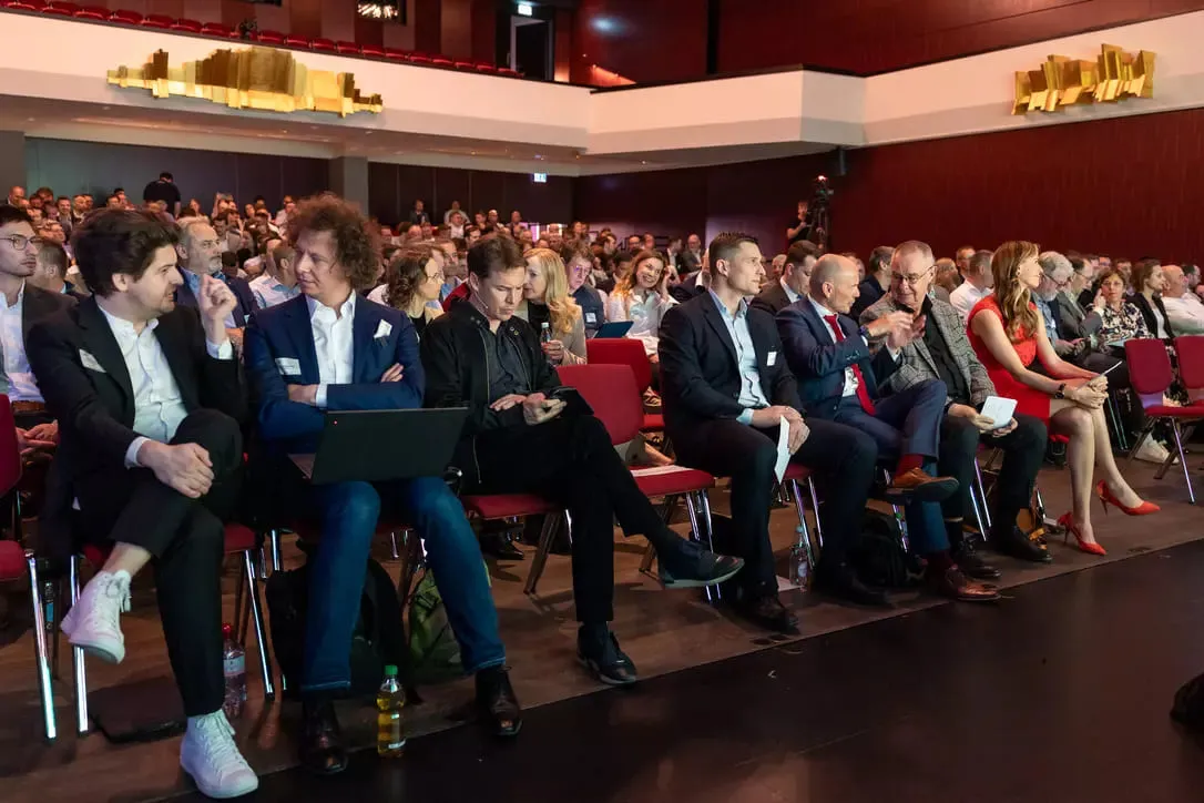 Audience seated in a conference hall, some engaged in conversation or using electronic devices.
