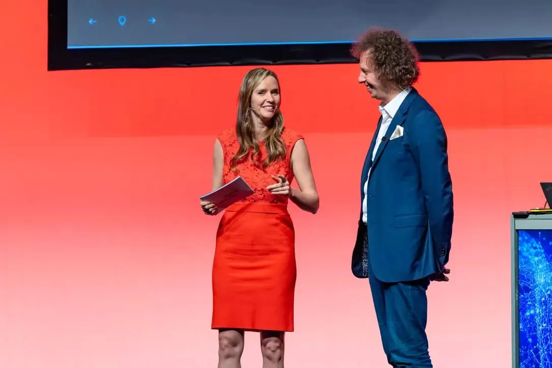 Woman in a red dress and man in a blue suit speaking on stage with a red background.