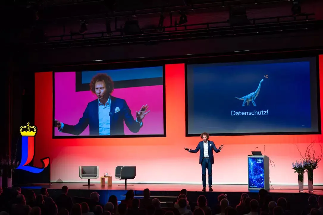 Man in a blue suit giving a presentation on stage with two large screens behind him; one screen shows his image and the other displays a dinosaur graphic with the word 'Datenschutz!'