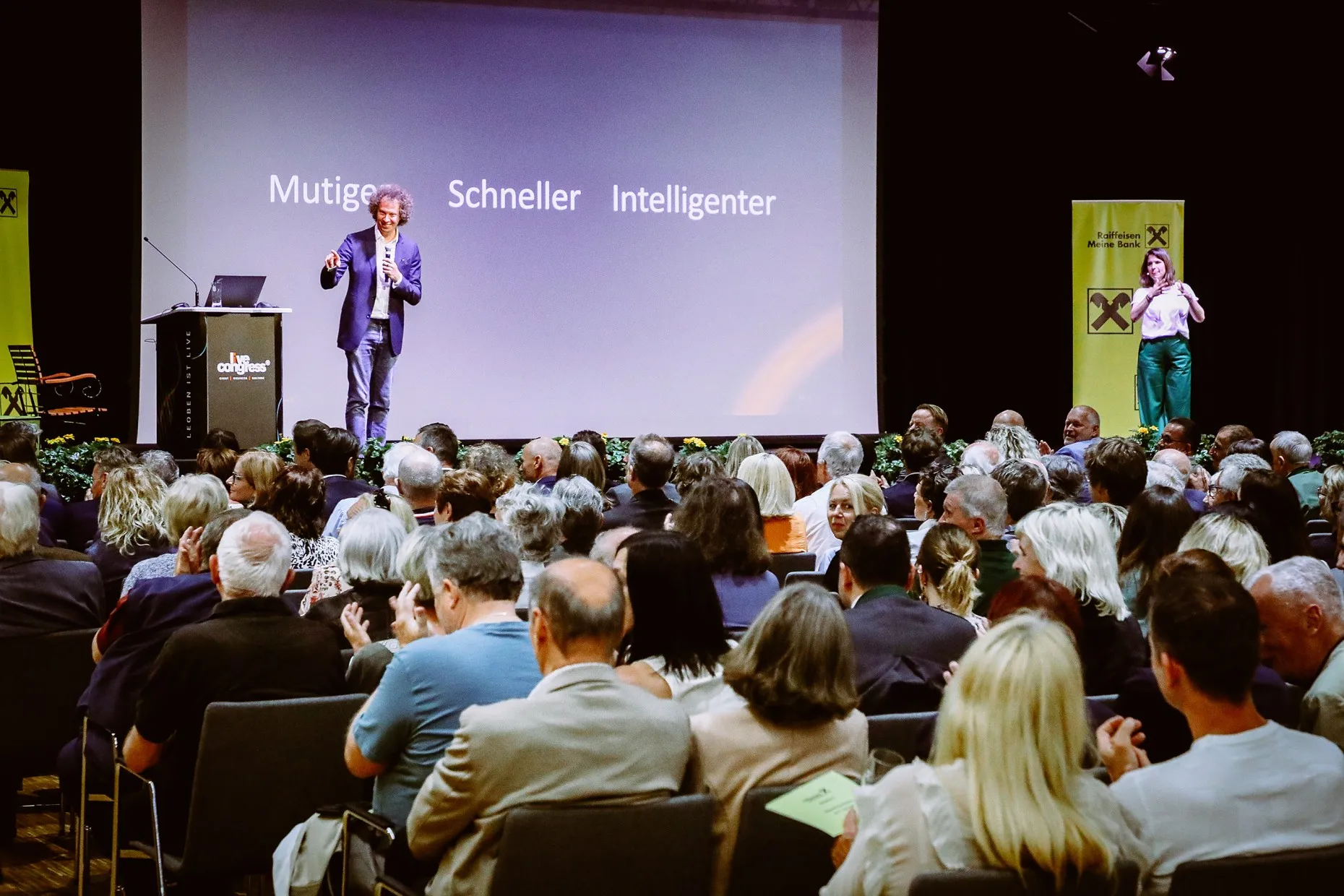Speaker presenting on stage in front of an audience with words Mutiger, Schneller, Intelligenter displayed on screen.