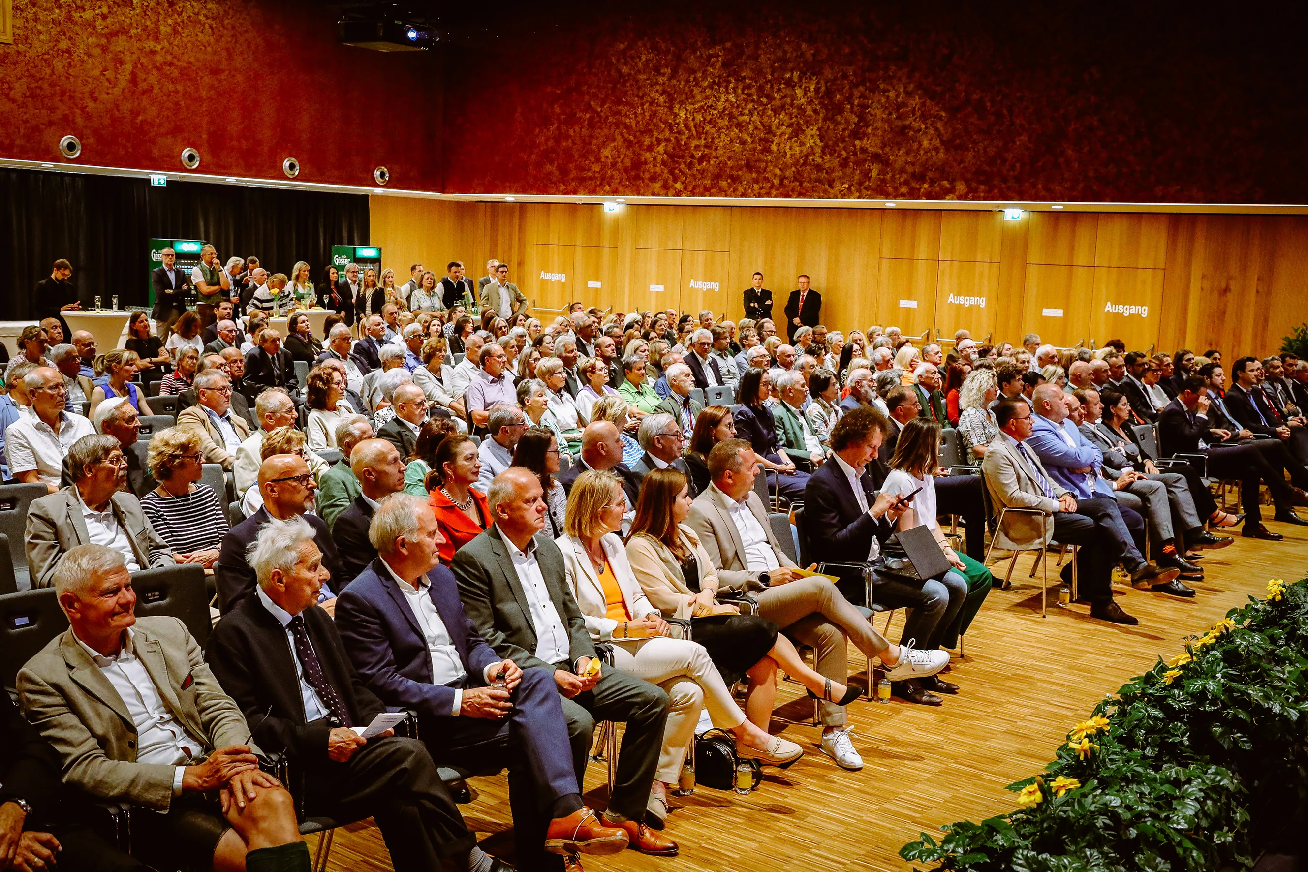 Large audience seated in a conference hall with wooden walls and a red textured upper wall, attentively listening to a speaker.