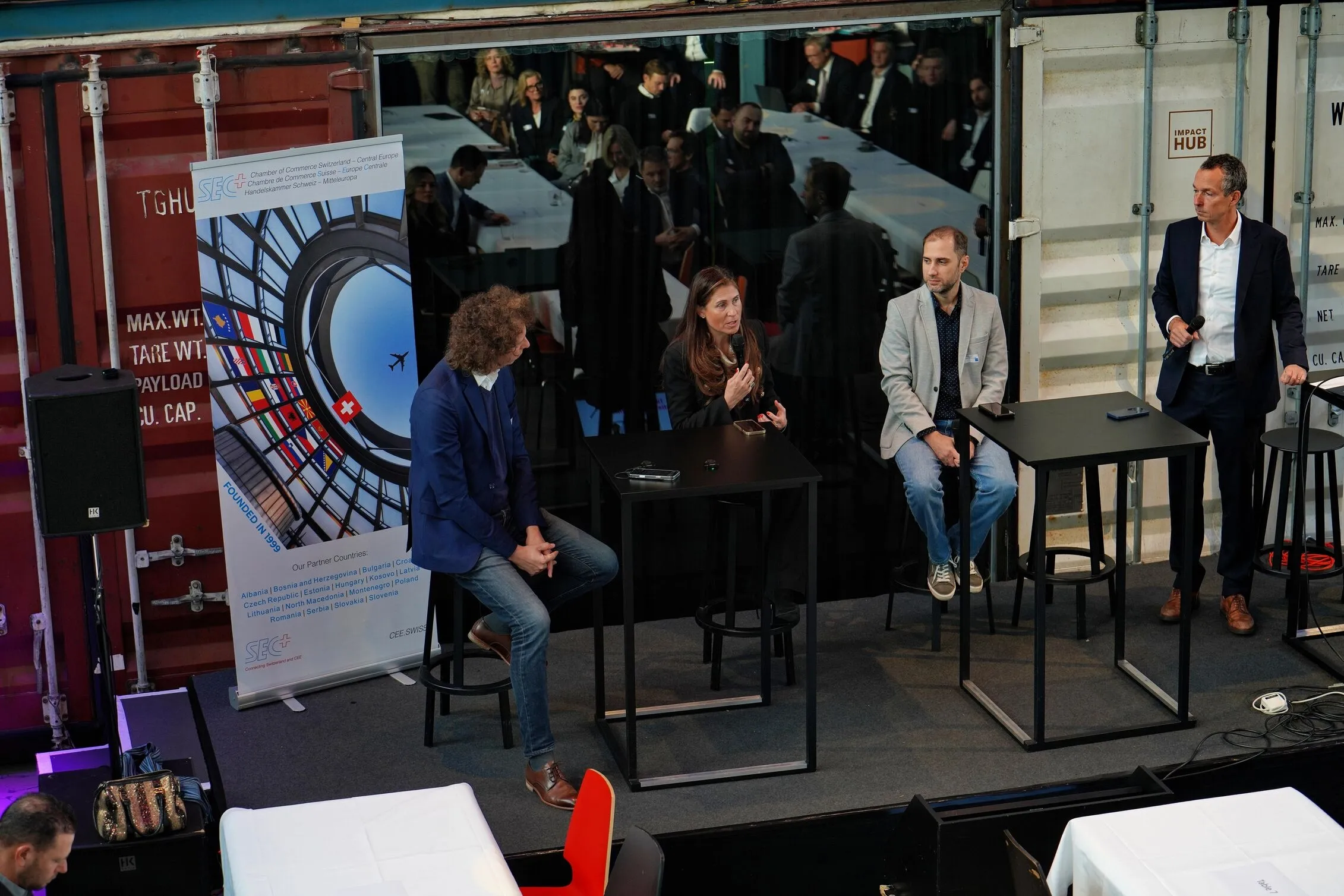 Four people engaged in a panel discussion on a stage with black tables and stools, a banner displaying European flags and Chamber of Commerce Switzerland text, and an audience reflected in a glass wall behind them.