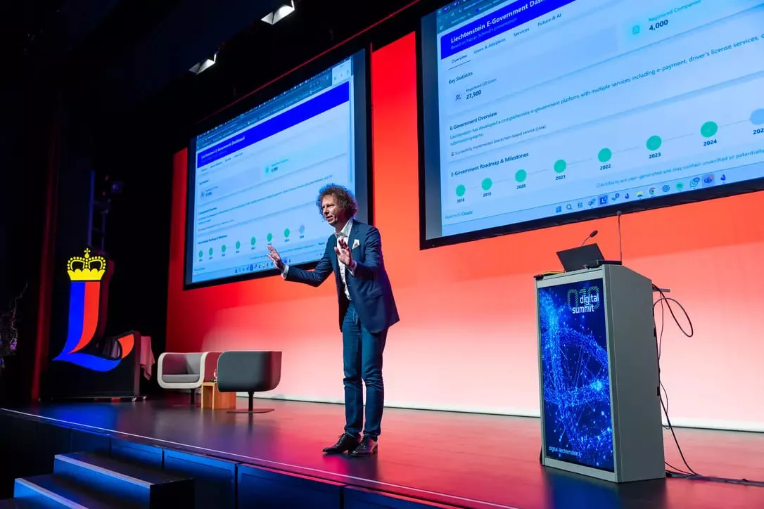 Presenter in a suit speaking on stage with two large screens displaying Liechtenstein E-Government dashboard and a podium labeled Digital Summit.