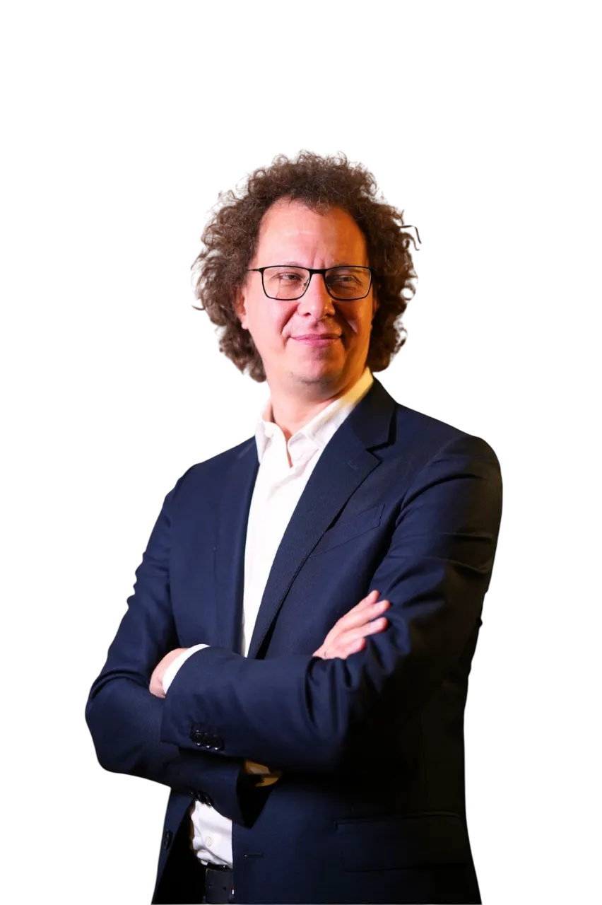 Man with curly hair and glasses wearing a dark suit and white shirt, standing with arms crossed and looking to the side.