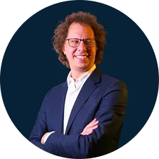Smiling man with curly hair and glasses wearing a navy blazer and white shirt with arms crossed.